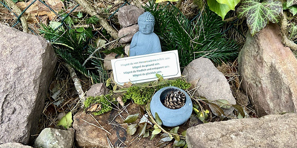 Buddha-Figur am Wegesrand mit Schälchen, Steinen, Moos, Blättern und Kärtchen mit Segensworten