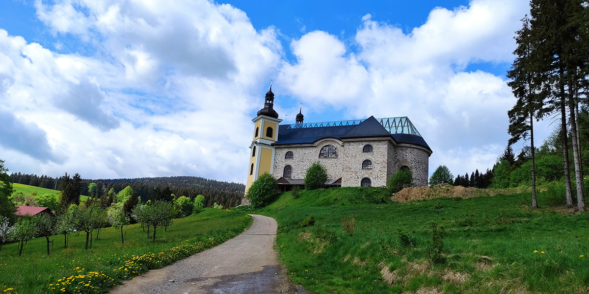 Ein Feldweg im Grünen Hügelland, der auf die Kirche Mariä Himmelfahrt zuführt; das Kirchendach besteht zum Teil aus Glas